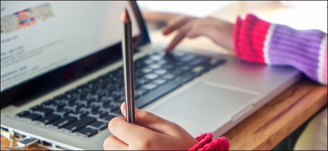 A child's hands typing on a MacBook and holding a pencil.