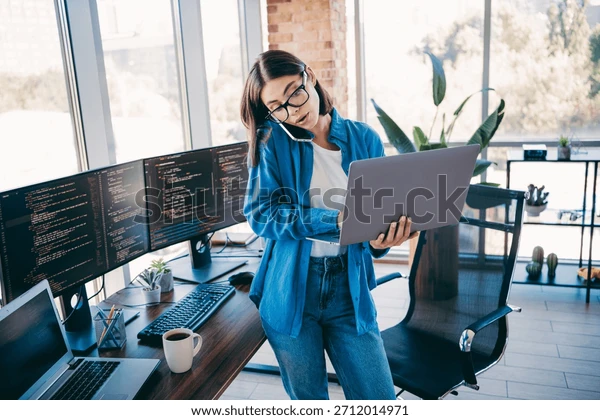 图片来源：一名职业女商人站在一间明亮的阁楼办公室里，一边拿着笔记本电脑，一边打电话，四周环绕着编程屏幕，演示技术工作、编程和远程协作。库存照片2712014971  | Shutterstock